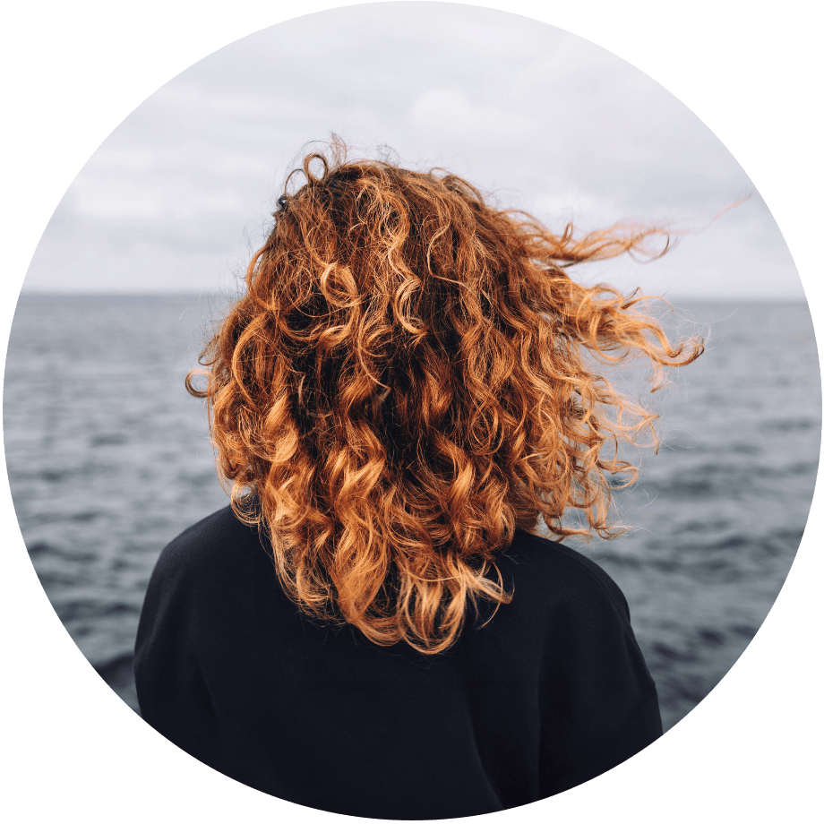 Eine dem Meer zugewandte Frau mit im Wind wehendem lockigem Haar.