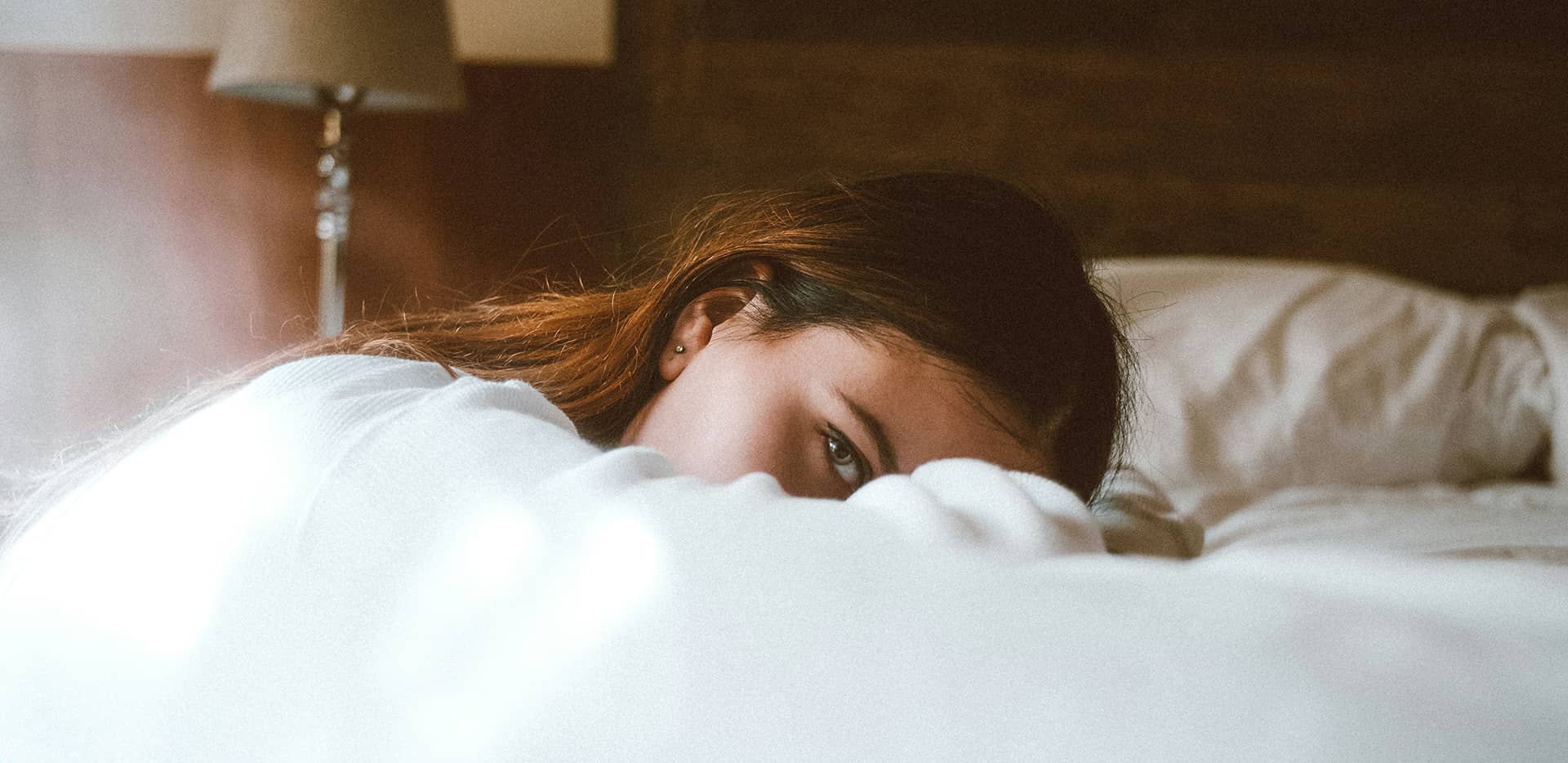 Eine junge Frau, die mit überkreuzten Armen auf dem Bett liegend den Betrachter sinnend anschaut.